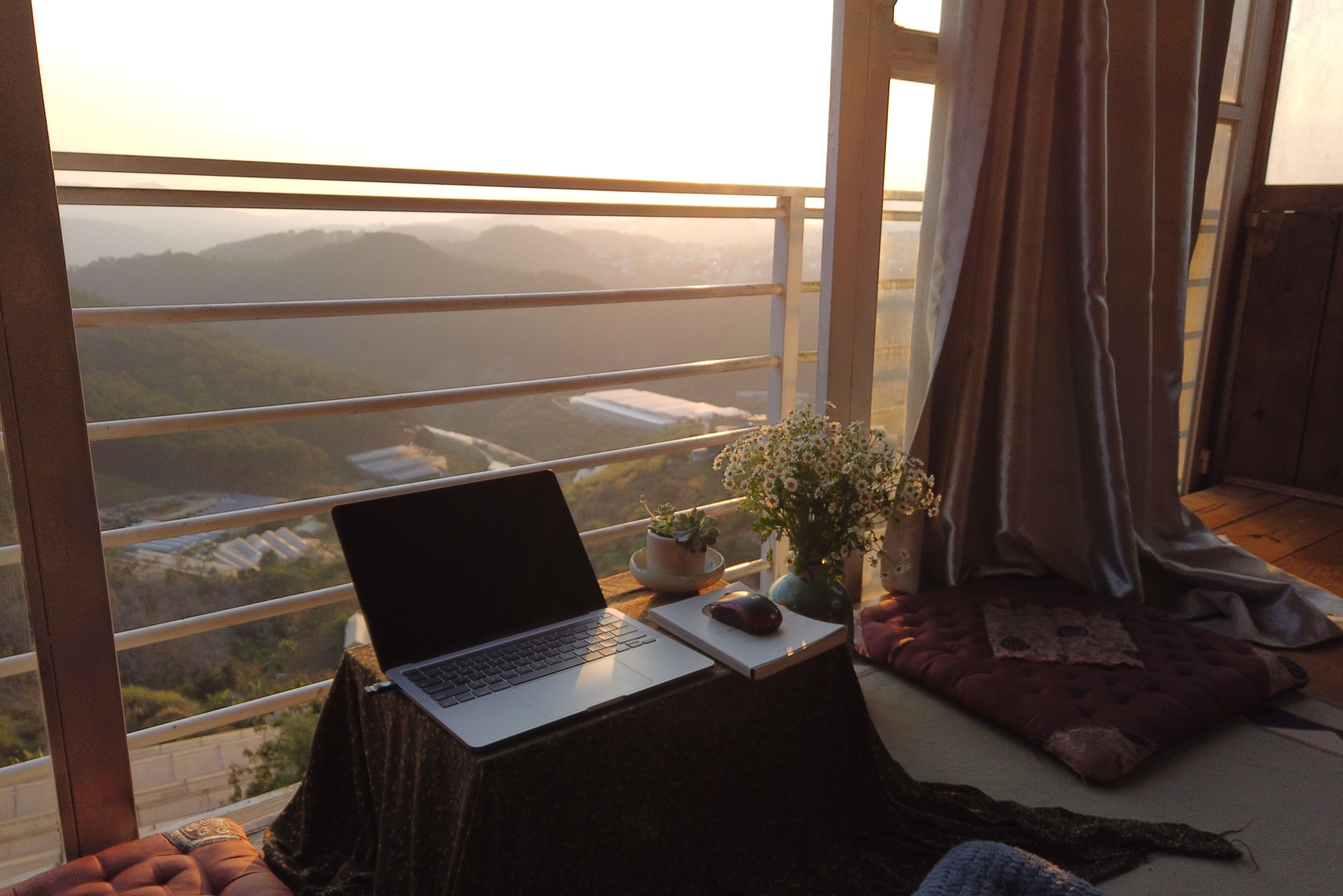 A serene workspace with a laptop and flowers overlooking Dalat's picturesque hills at sunset.