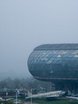 Futuristic glass building amidst fog with aircraft displayed outdoors.