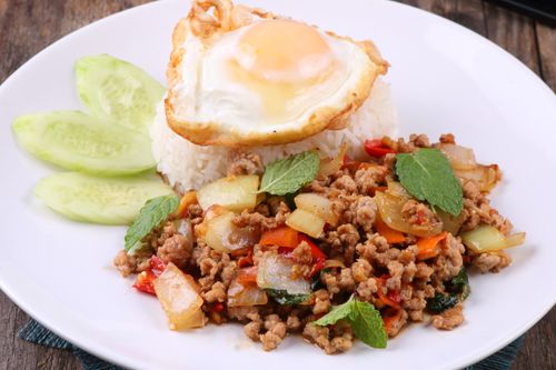 Free Authentic Thai basil pork with rice, fried egg, and garnished cucumbers on a white plate. Stock Photo