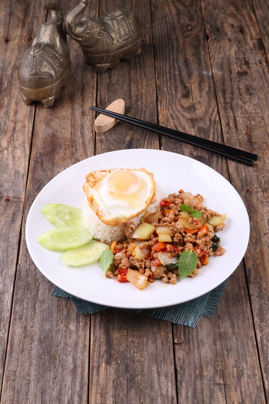 Bowl of spicy Thai ground pork stir-fry topped with a fried egg, served over jasmine rice with wilted basil visible throughout