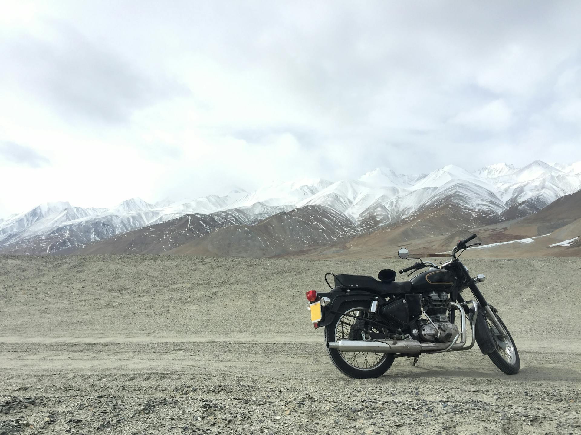 A vintage motorcycle parked on a gravel road with snowcapped mountains in the background, under a cloudy sky.