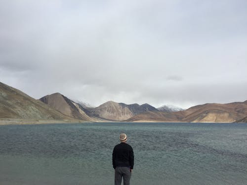 Person Standing Facing Body of Water With Hills at Distance