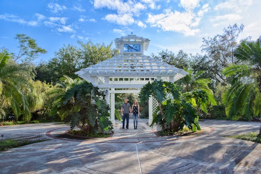 A couple stands under a serene pergola surrounded by lush greenery on a sunny day.