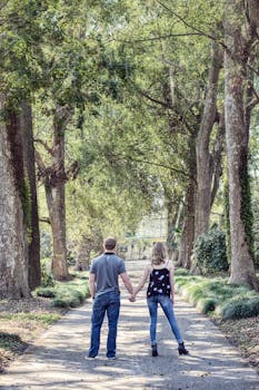 A couple holds hands while walking down a picturesque tree-lined path surrounded by nature.