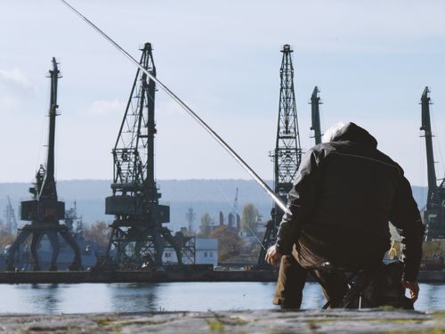 Free A person fishing by the waterfront with industrial cranes in the background. Stock Photo