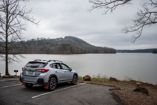 Silver SUV in a lakeside parking lot with mountain and tree view under cloudy sky.