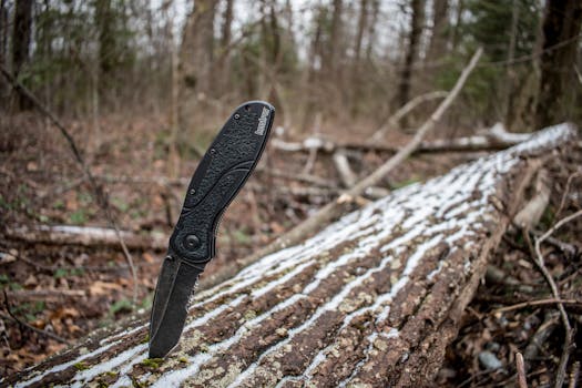 Close-up of a tactical folding knife embedded in a fallen log within a forest, evoking outdoor adventure.