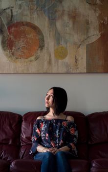 Thoughtful woman sits on sofa, gazing at abstract painting above.