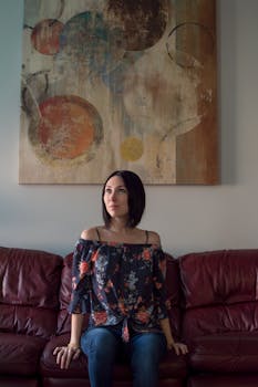 A woman sits thoughtfully on a leather couch beneath abstract wall art.