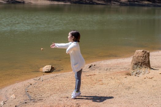 A child in a white jacket throws a rock by a peaceful lakeside on a sunny day, enjoying outdoor activities.