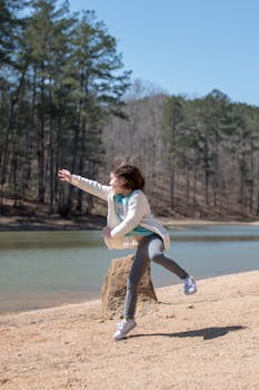 A young girl joyfully plays near a serene river, surrounded by lush woodland, under a clear blue sky.