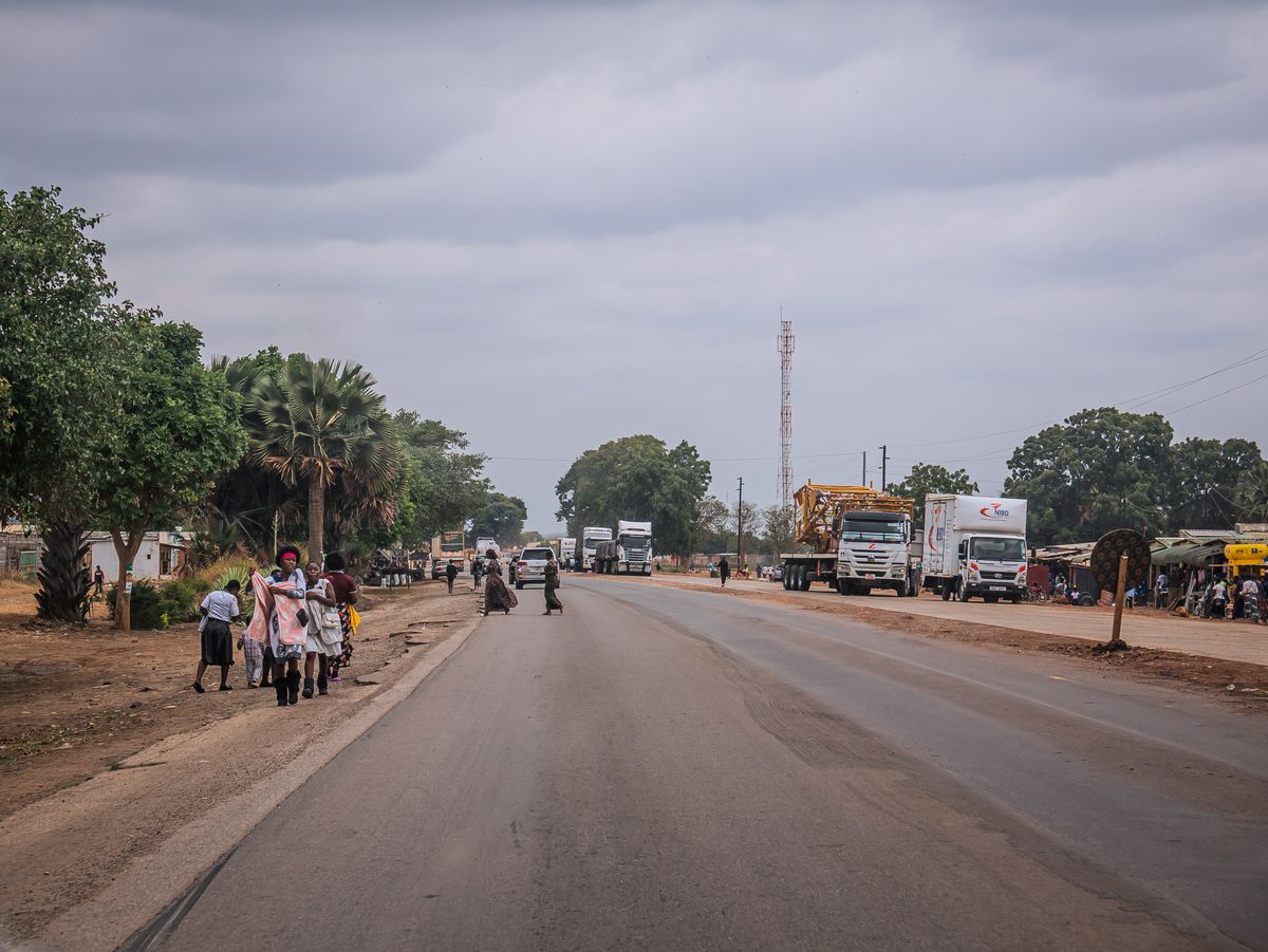 A roadside scene in an African town with pedestrians, parked and moving trucks, and shops lining the road under a cloudy sky
