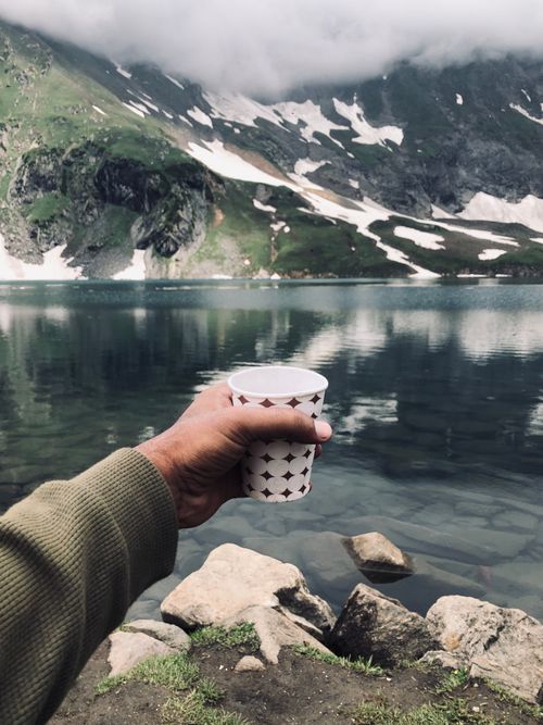 Free A person holds a cup by a tranquil lake with snow-capped mountains in view. Stock Photo