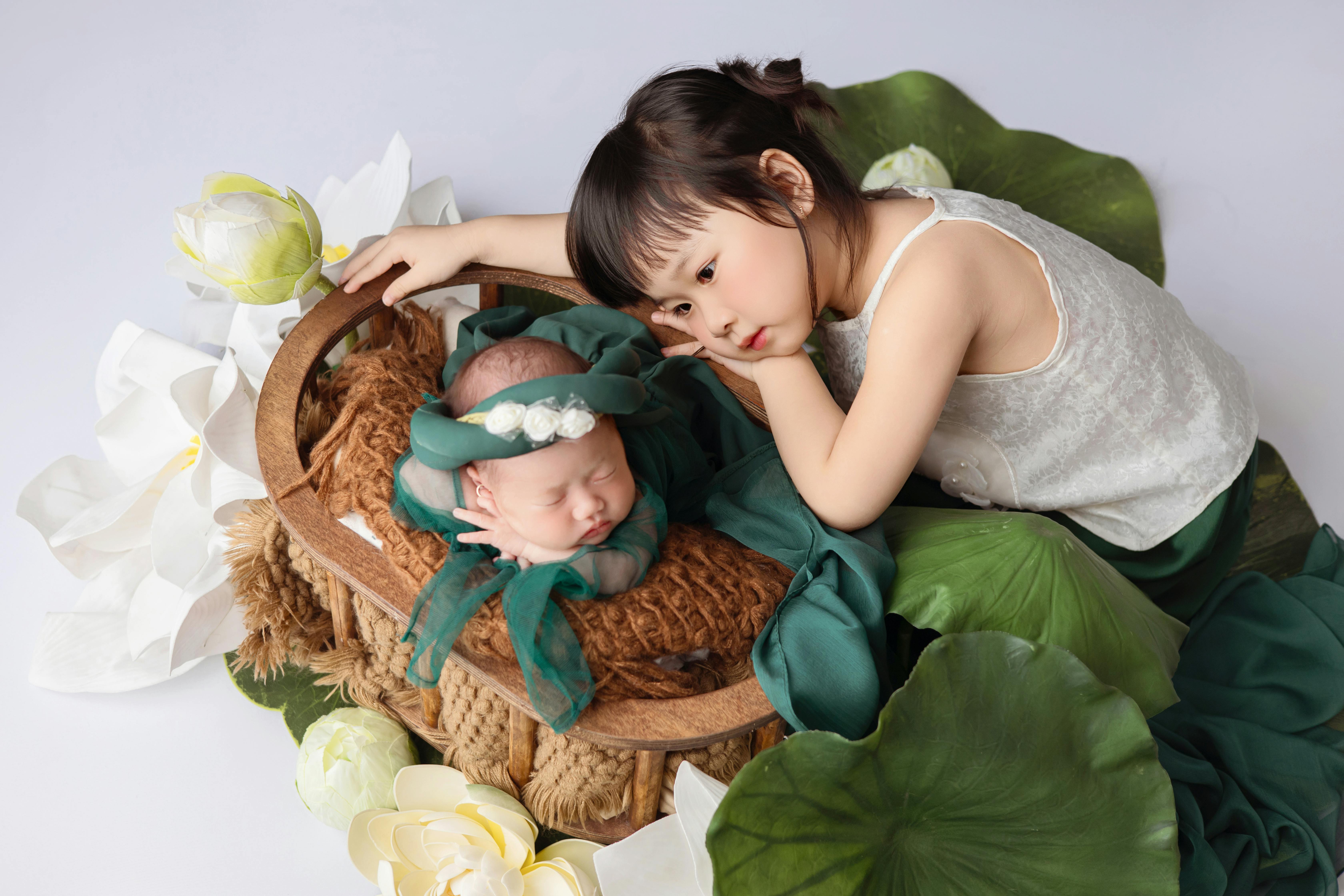 A touching scene of a sibling watching over a sleeping newborn in a lotus adorned nest.
