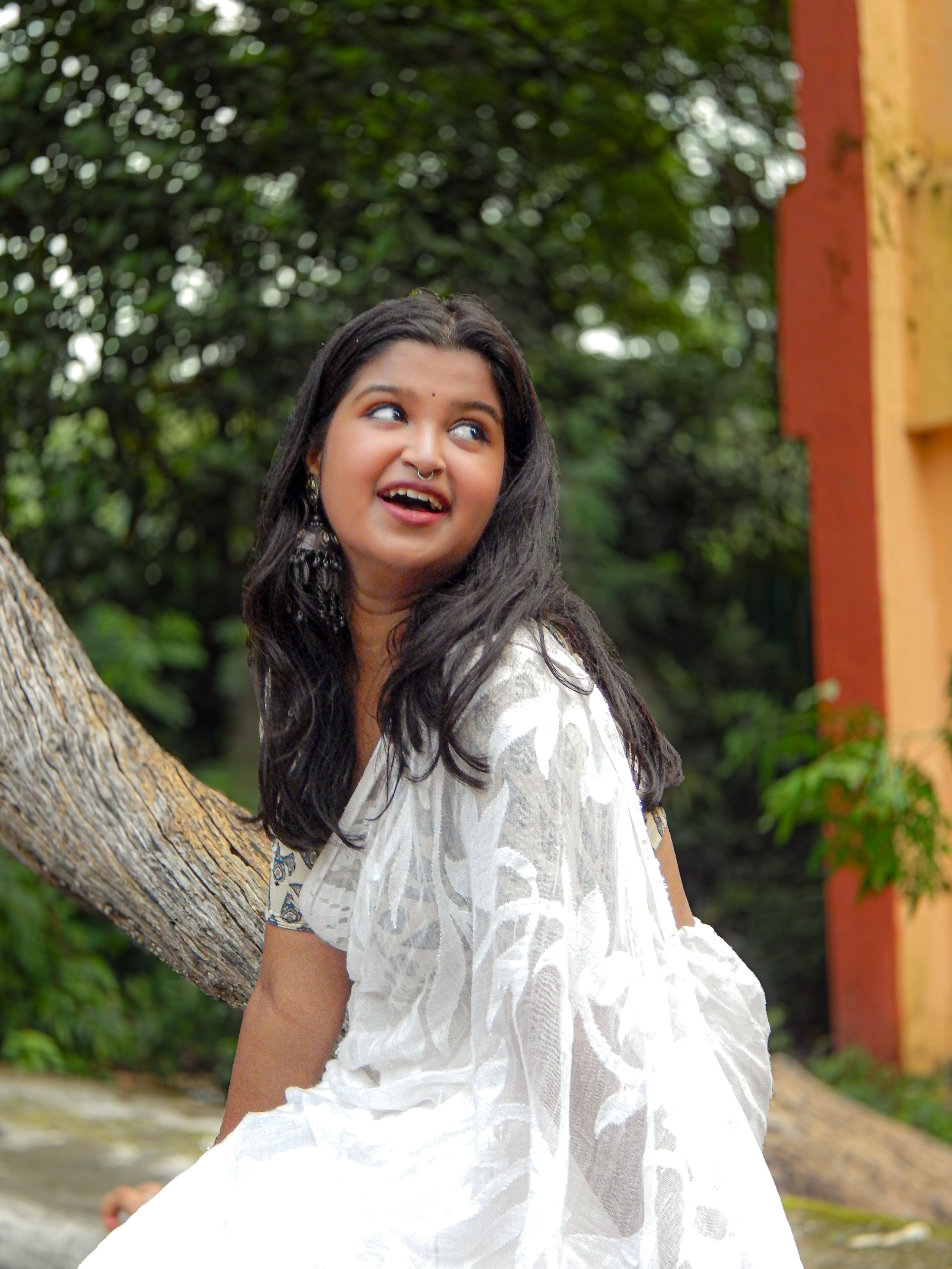 Woman in a white saree sitting on a tree, smiling and enjoying the outdoor scenery.