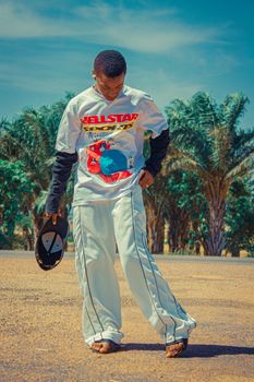 African teenage boy in casual fashion, posing outdoors with vibrant palm trees and blue sky.