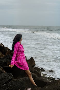 Woman in a pink dress sitting on rocks by the ocean, staring at the waves.