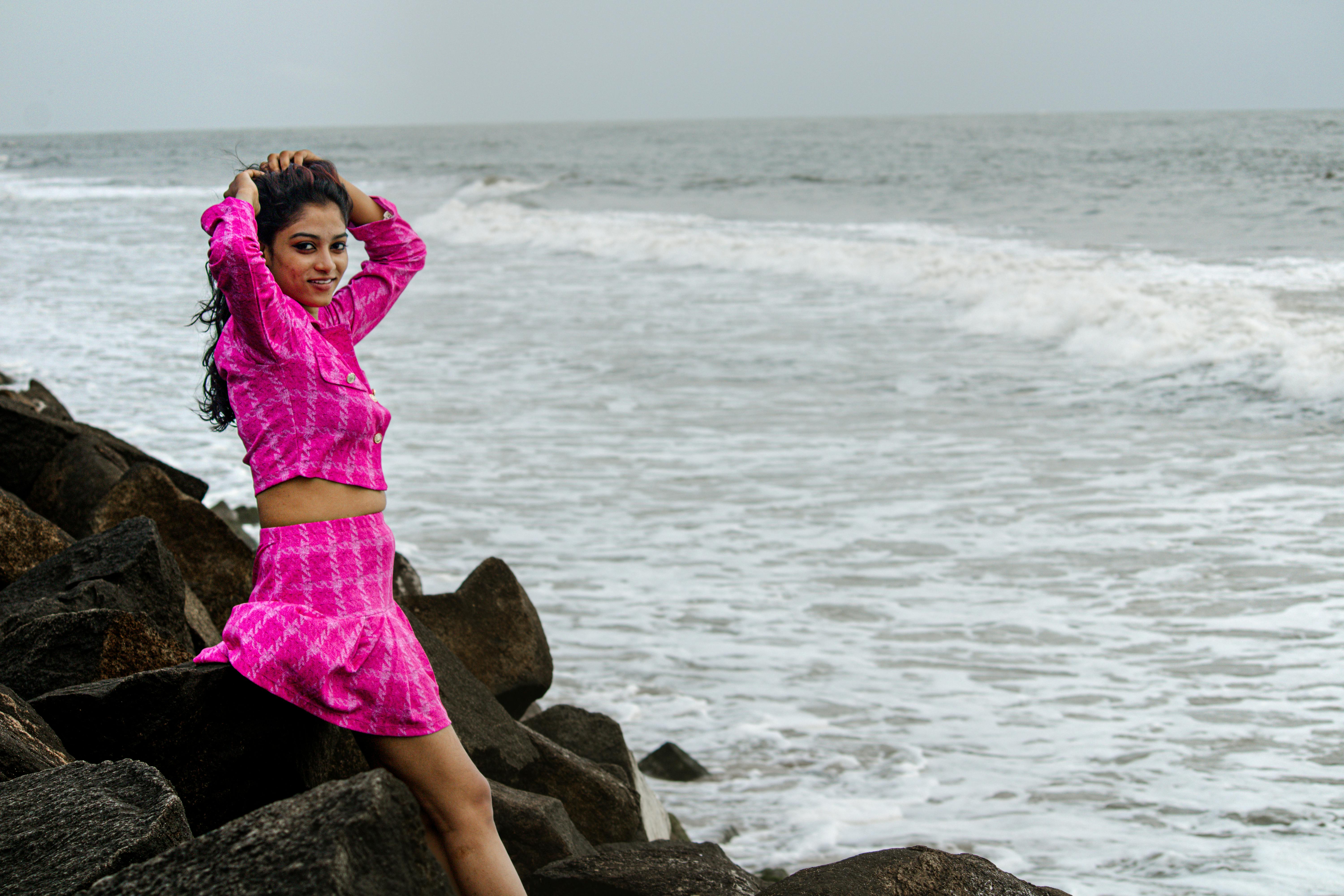 Woman in pink outfit posing on rocks by the ocean, exuding confidence and style.