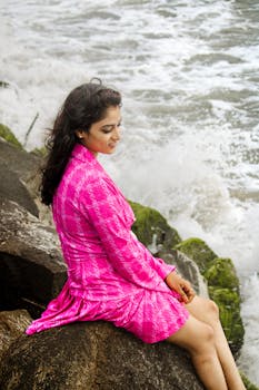 A woman in a vibrant pink dress sits on rocks beside the ocean, enjoying a serene moment.
