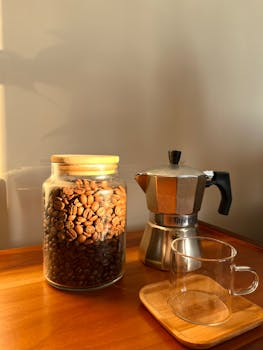 A cozy setup featuring a jar of coffee beans, Moka pot, and glass cup in warm lighting.