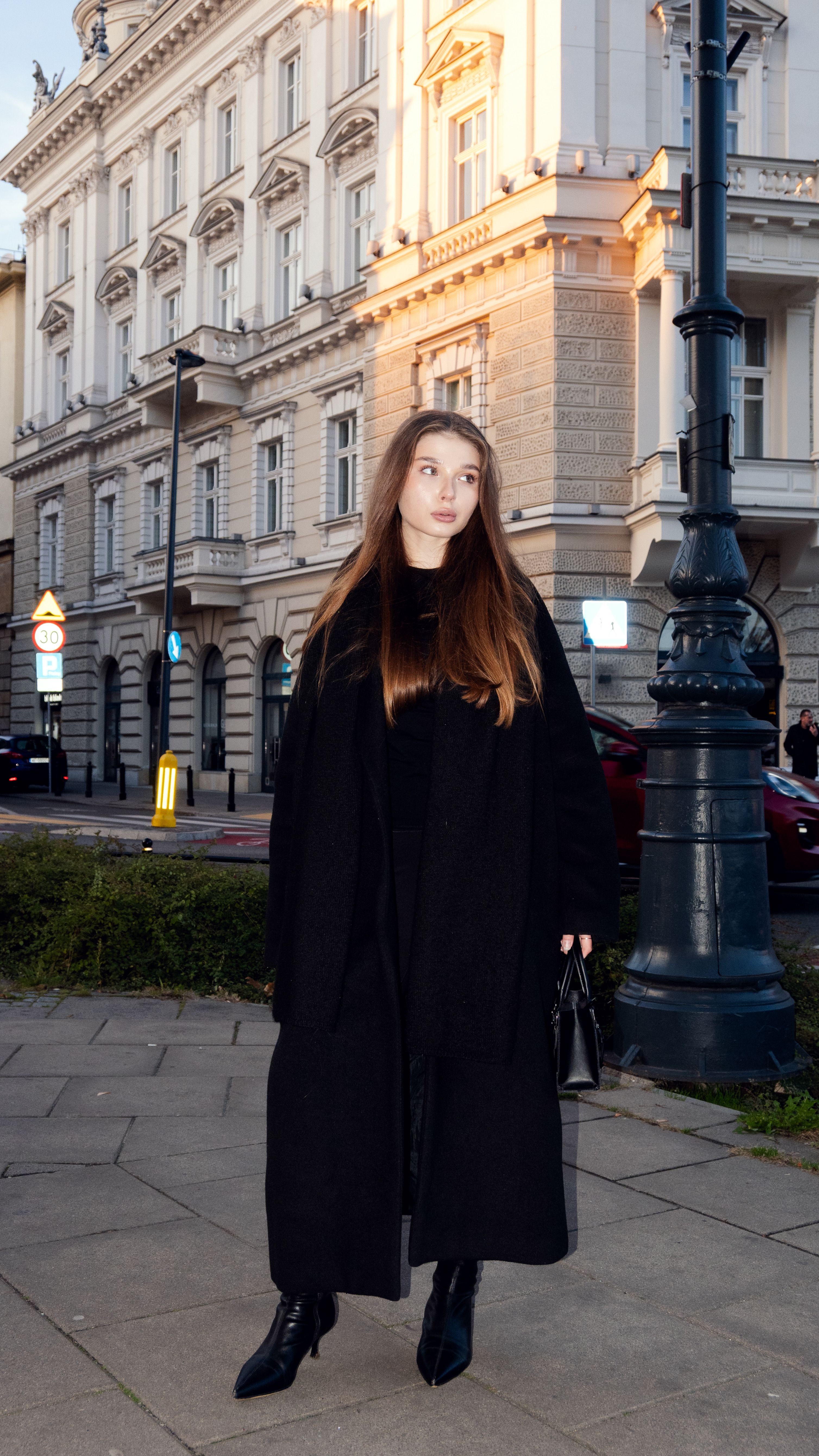 Stylish woman in black coat stands confidently in front of historic urban architecture.