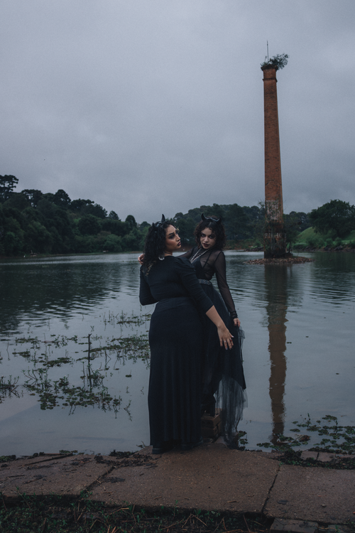 Free Two women in gothic attire pose by a lake with a towering column in the background. Stock Photo