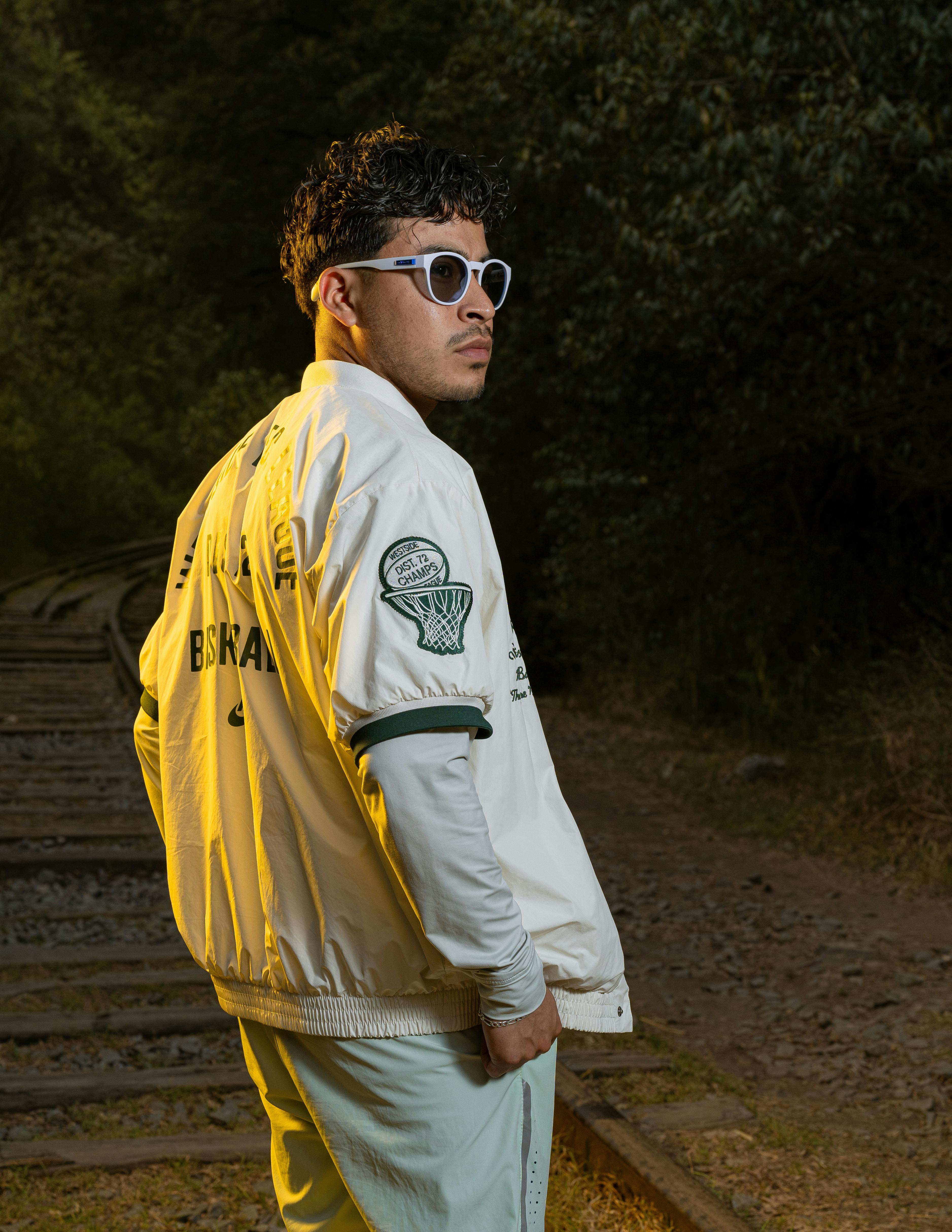 Fashionable man with sunglasses and jacket poses confidently on railway tracks at dusk.