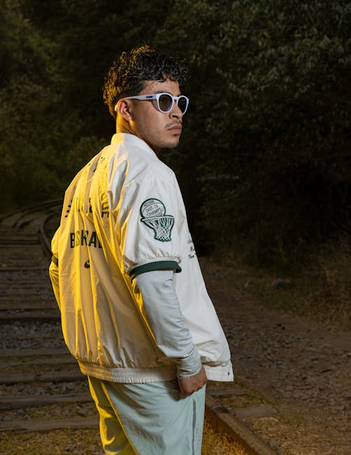 Fashionable man with sunglasses and jacket poses confidently on railway tracks at dusk.