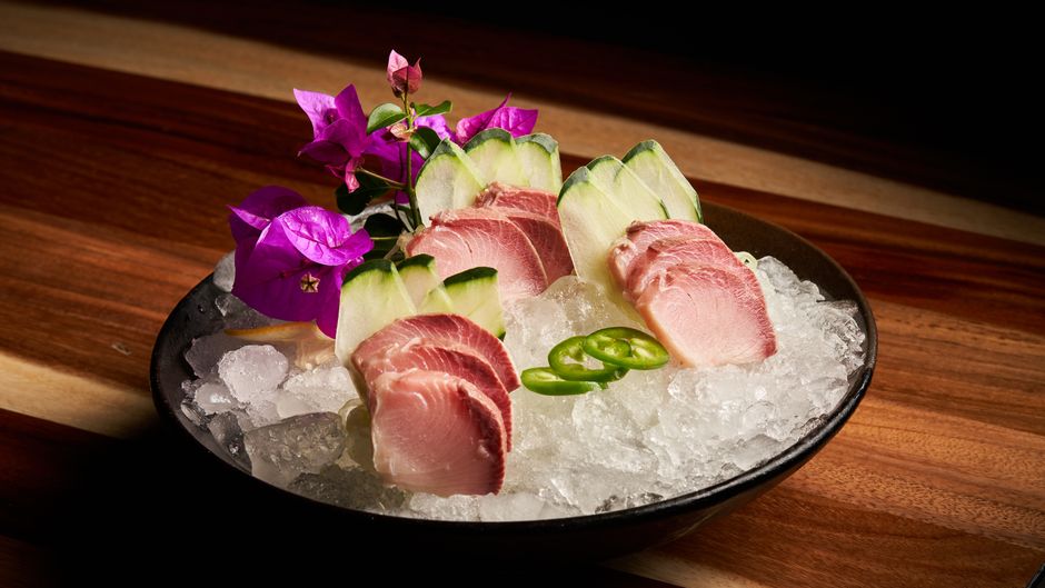 Thin slices of pale pink hamachi arranged on a white plate, drizzled with olive oil and topped with sliced jalapeño and citrus zest