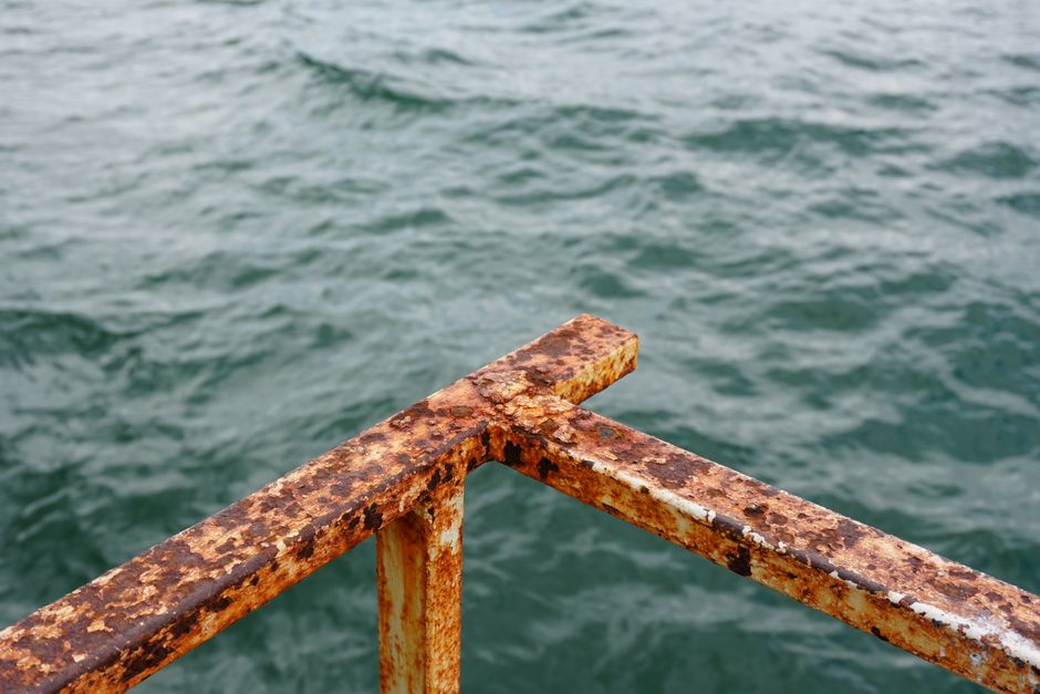 Close-up of a rusted metal railing with ocean waves in the background, suggesting decay and nature's force.