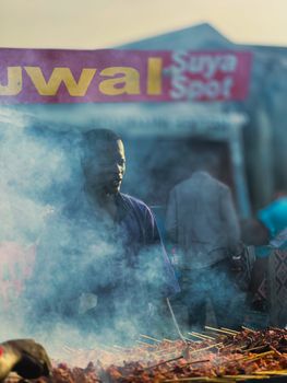 A street vendor grills skewers at a smoky outdoor market scene, with visible signage in background.