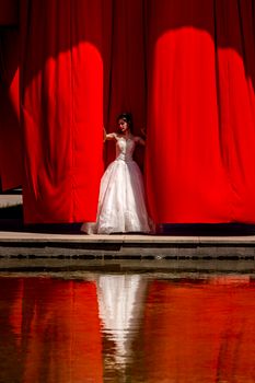 A bride stands against a vibrant red curtain, her reflection visible in a calm pool below.