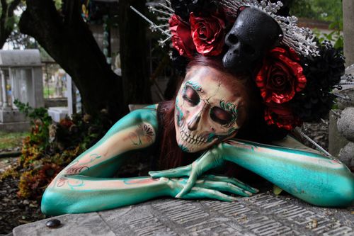 Free A person in vibrant catrina makeup and ornate headdress with red roses, lying in a somber cemetery setting. Stock Photo