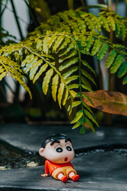 A small cartoon figurine sits under vibrant green ferns on a stone surface.