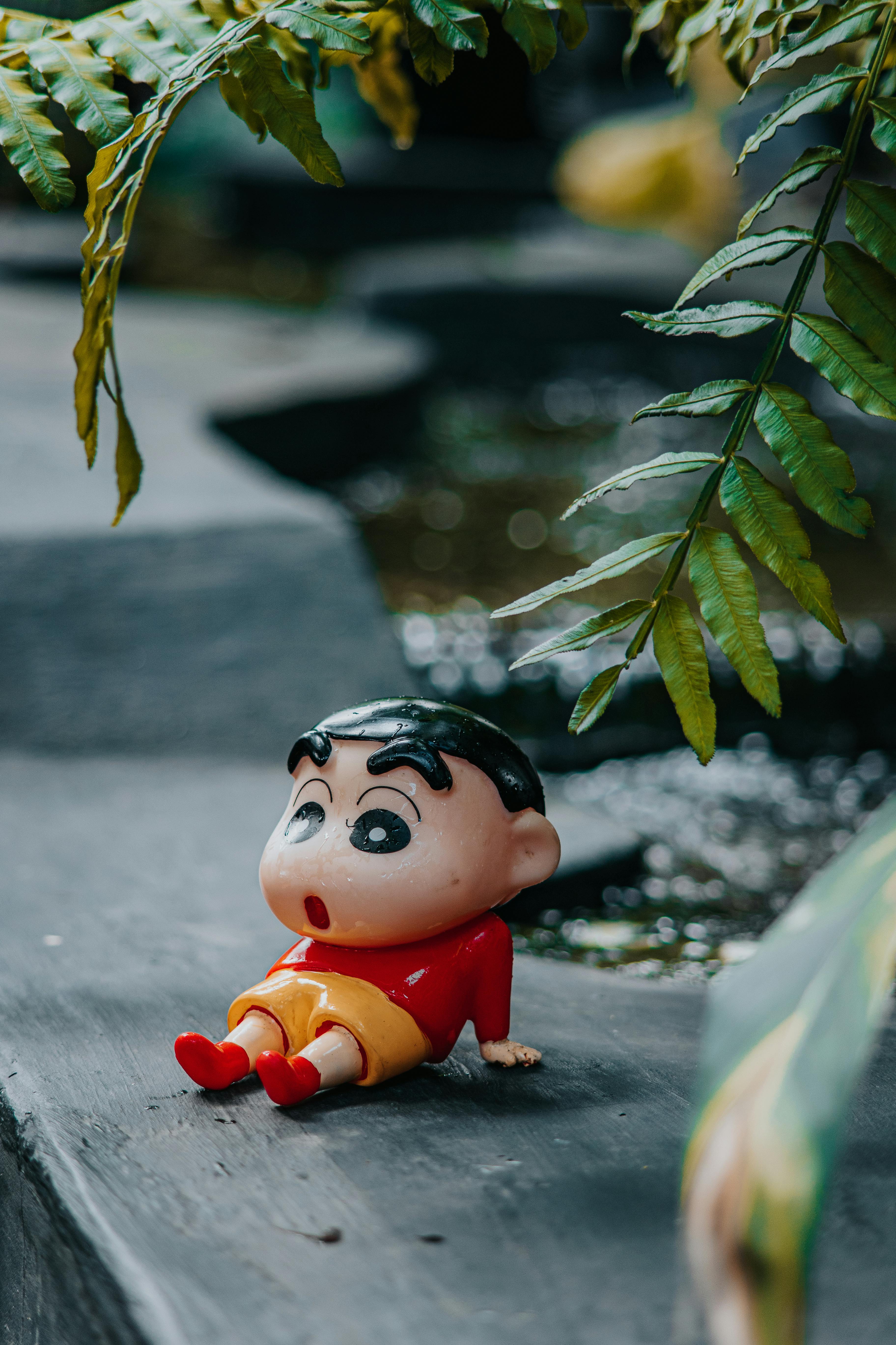A vibrant cartoon character figurine rests on a dark stone, surrounded by lush green plants.