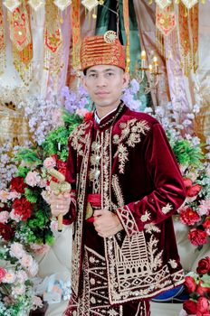 Portrait of a groom in traditional ornate red attire surrounded by vibrant flowers - Fotografer Sidoarjo