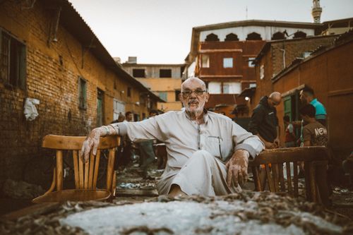Free Elderly man seated outdoors in a vibrant Egyptian street, surrounded by urban life. Stock Photo