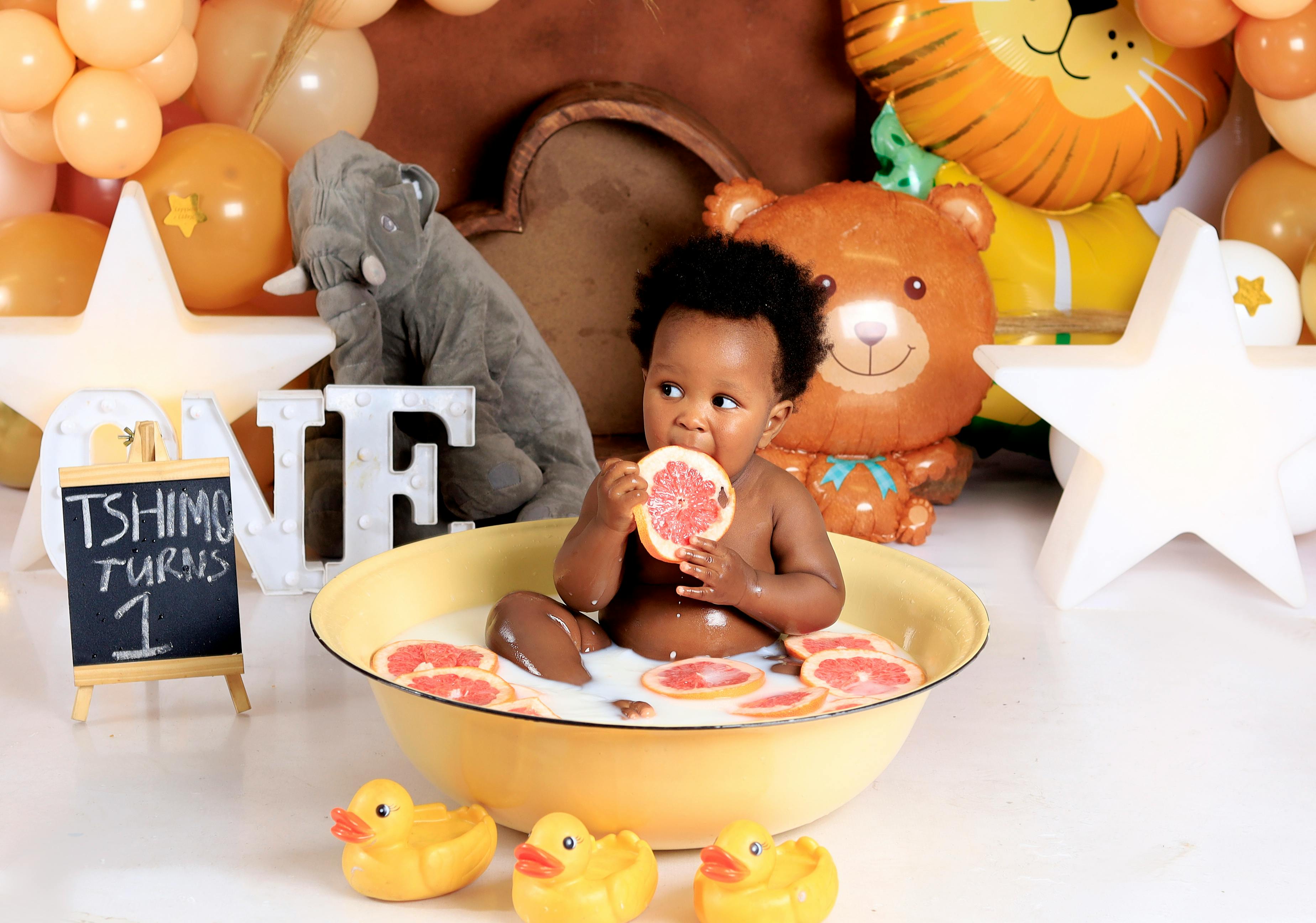 Cute baby boy enjoying a grapefruit bath on his first birthday surrounded by toys and balloons.