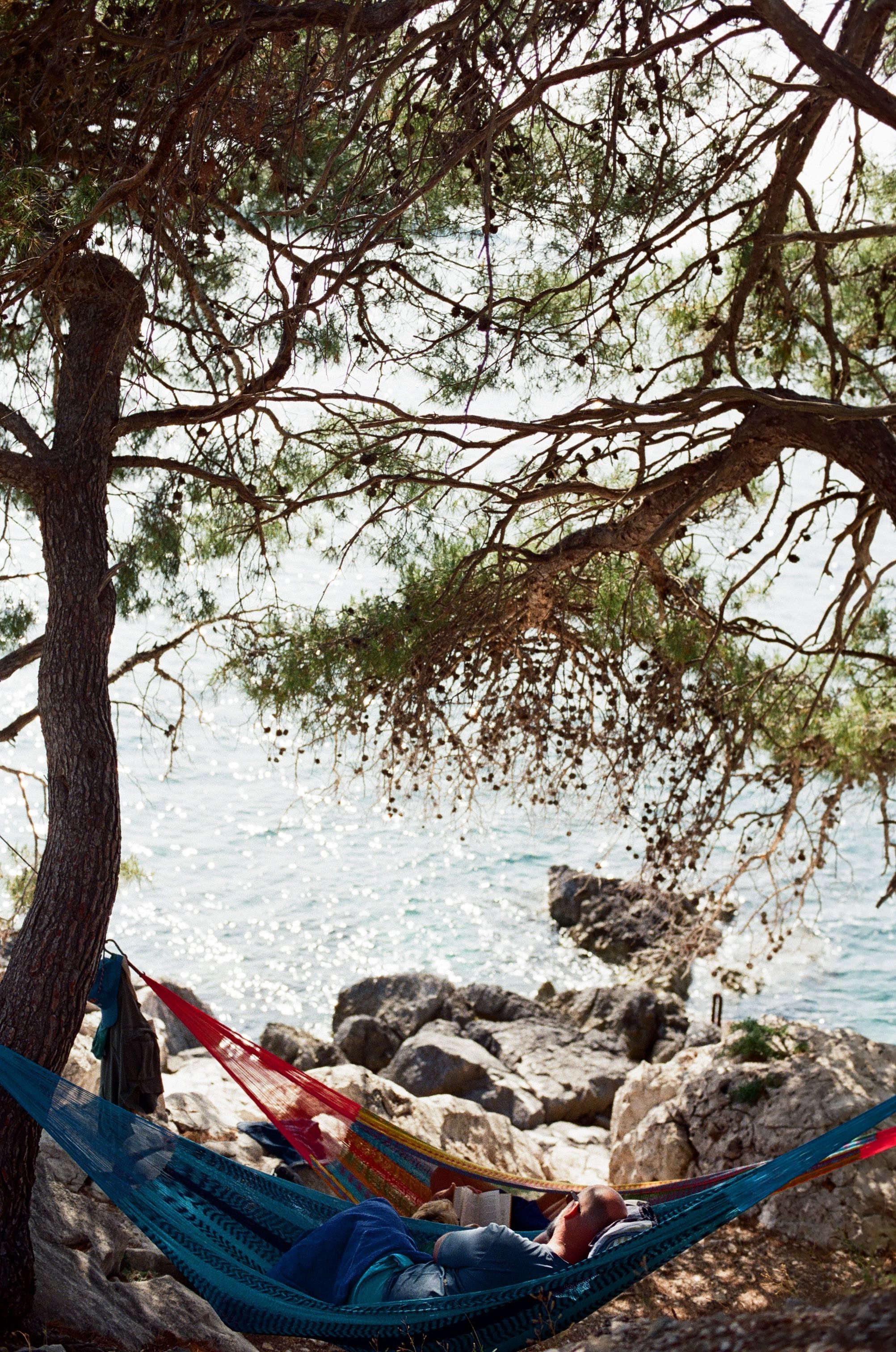 Person relaxing in a hammock under trees by a rocky seaside. Peaceful summer scene.