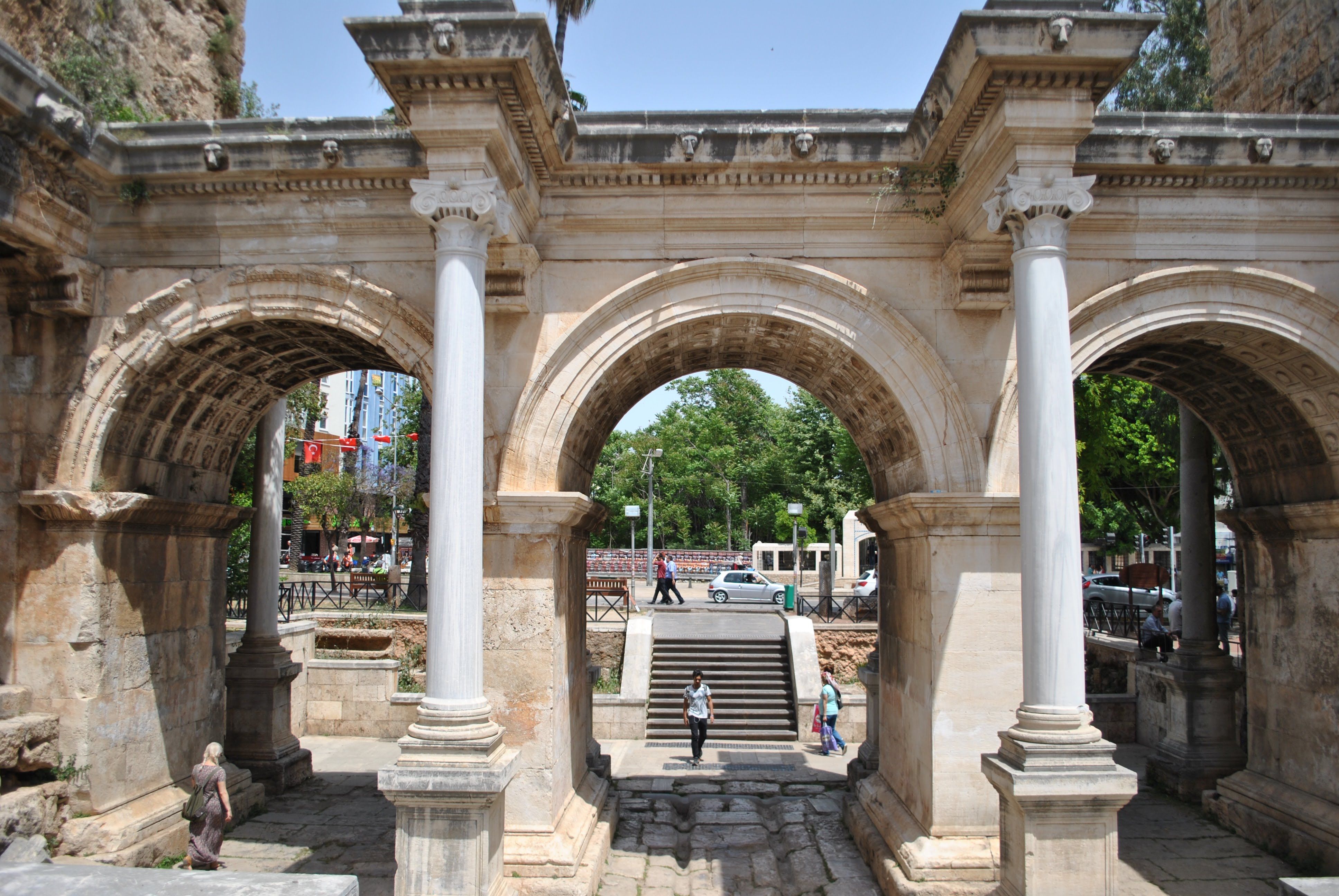 Front view of Hadrian's Gate in Antalya, Turkey, with visitors passing through the ancient Roman arches in bright sun.