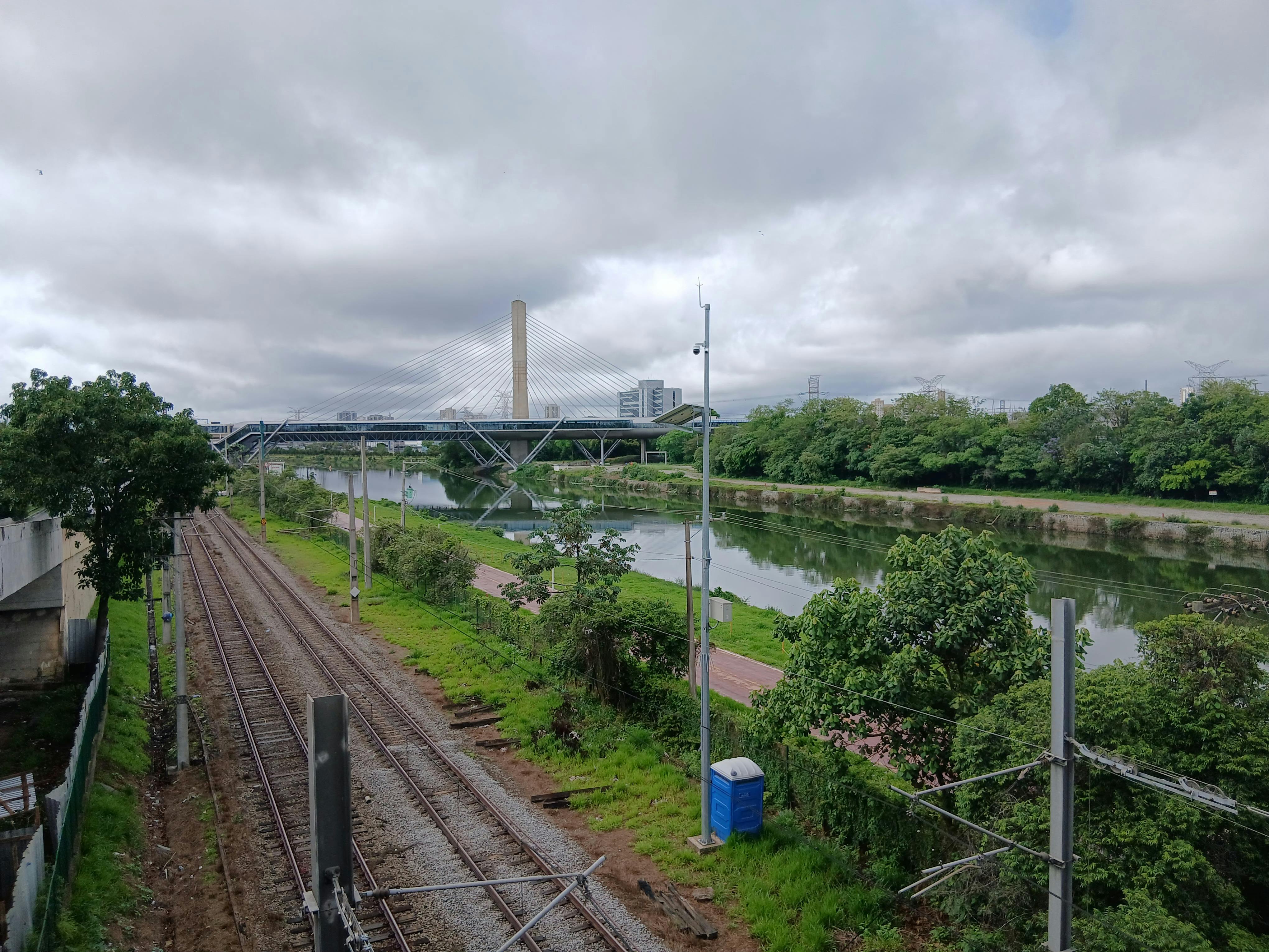 View of a modern bridge with railway tracks beside a river, lush greenery, and cloudy sky.