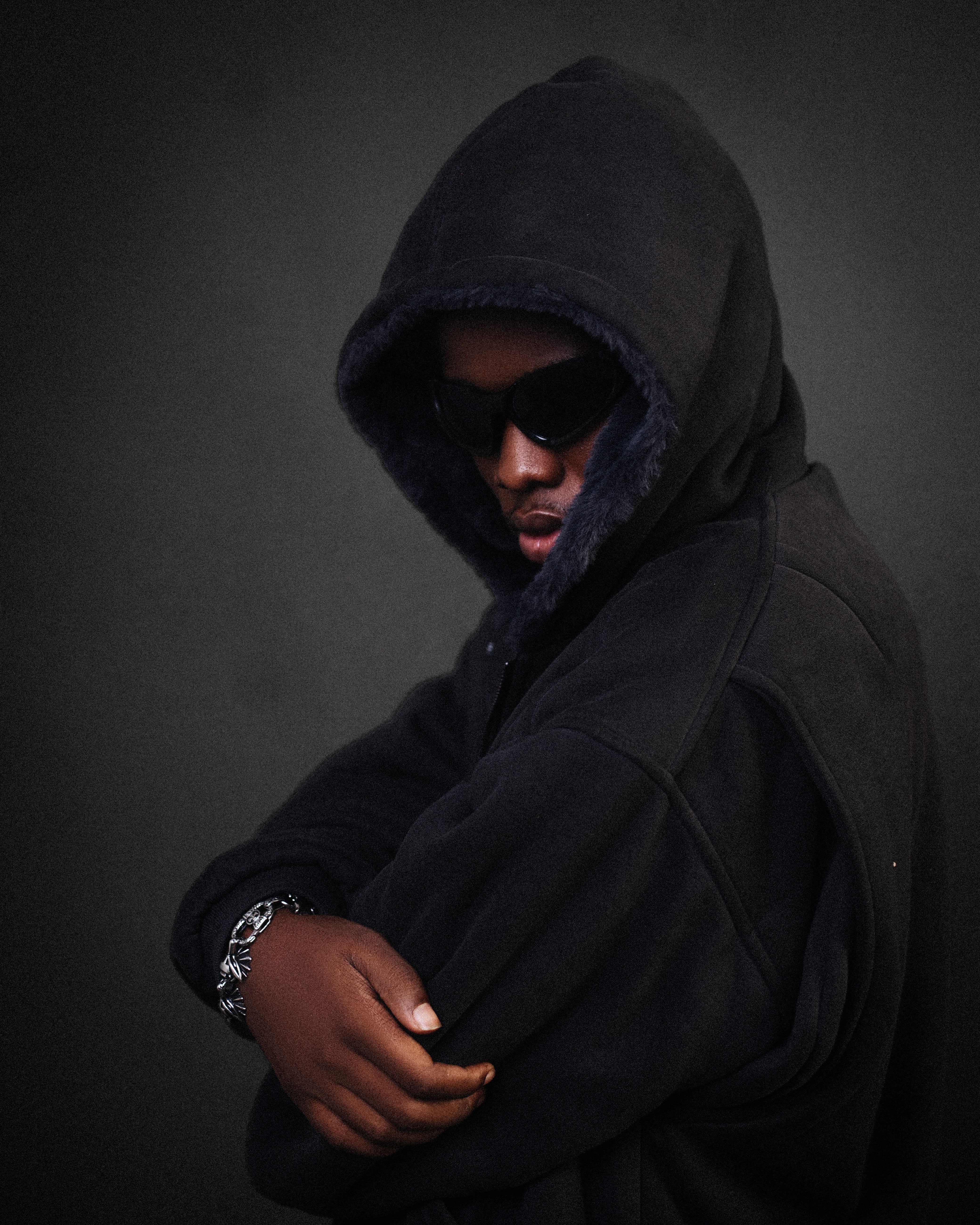 Portrait of a young man wearing a hoodie and sunglasses against a dark background.