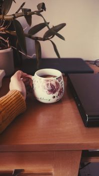 A comforting cup of tea held near a laptop in a cozy home office scene.