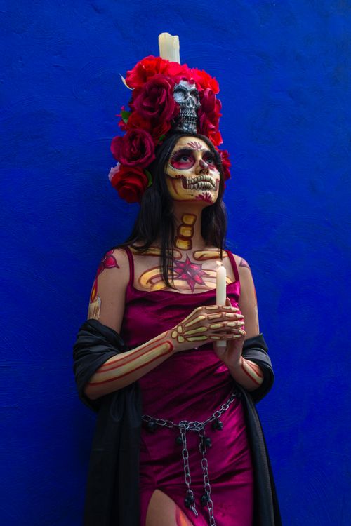 Free A woman in a colorful Catrina costume with floral headdress poses against a vivid blue wall. Stock Photo