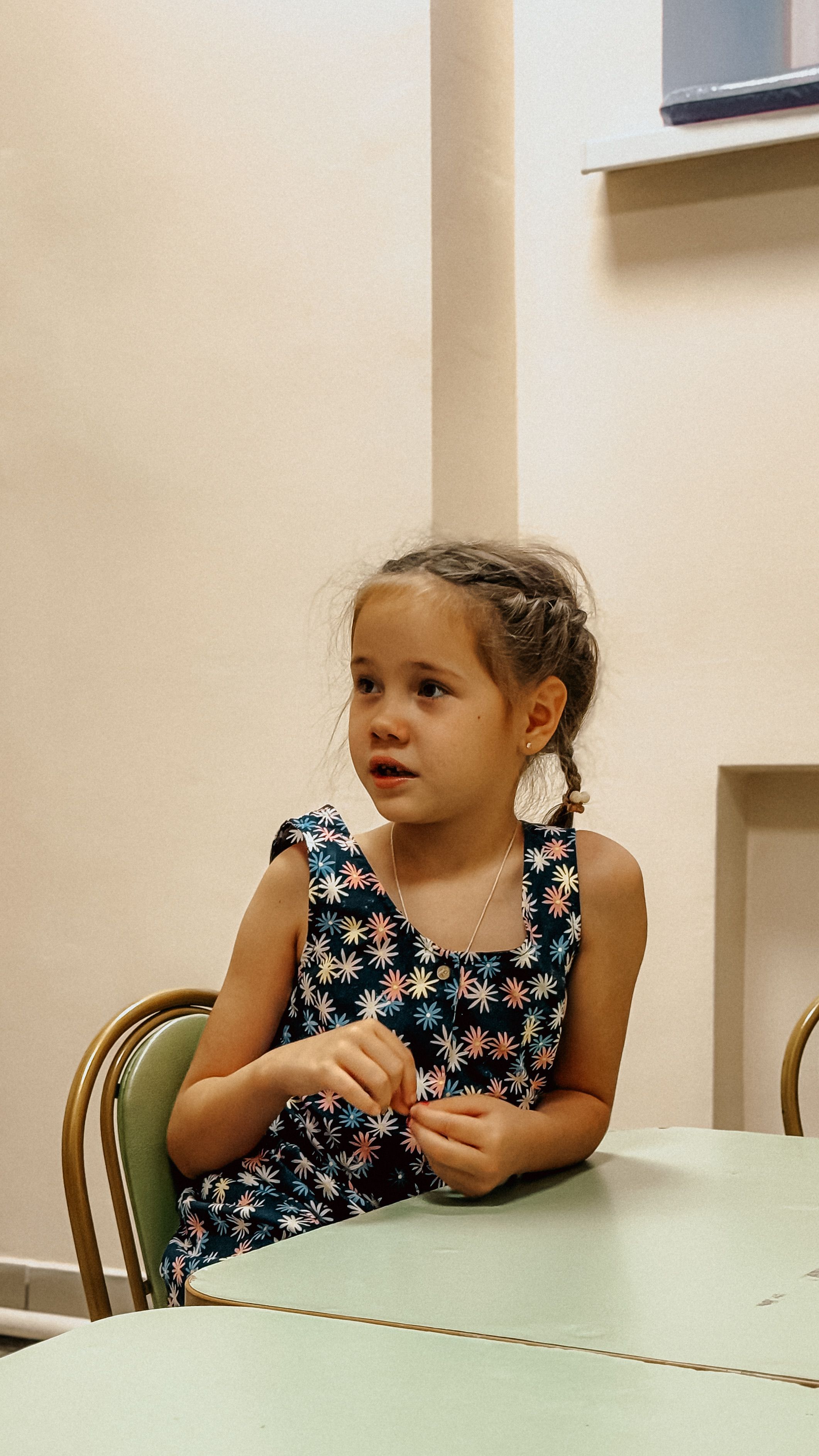 A young girl seated indoors, looking attentive in a classroom.