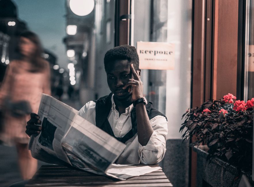 A fashionable man reads a newspaper at an outdoor cafe in the city at twilight, adding a touch of style to the urban scene.