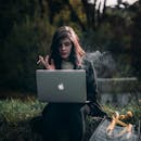 一位时尚女性在户外一边抽烟，一边用笔记本电脑工作，周围是大自然