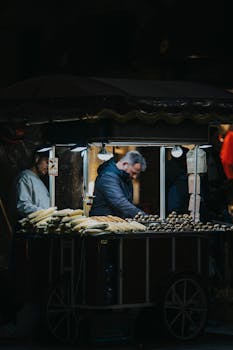 A street vendor serves grilled corn from a cart under night lights.