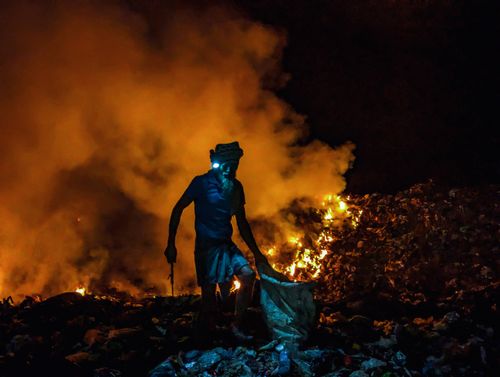 Free A silhouette of a person working amidst burning garbage piles at night, creating a dramatic scene. Stock Photo