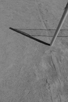 A monochrome image of a volleyball net casting shadows on a sandy beach.